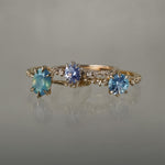 A group of three one of a kind rings by Laurie Fleming Jewellery. From left to right: An Ilona ring featuring an oval cut teal opalescent sapphire, a Nereid ring featuring an oval cut light blue purple pastel sapphire, and a Nereid ring with a John Dyer Starbrite cut blue sapphire centre. The rings are against a grey background.