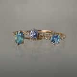 A group of three one of a kind rings by Laurie Fleming Jewellery. From left to right: An Ilona ring featuring an oval cut teal opalescent sapphire, a Nereid ring featuring an oval cut light blue purple pastel sapphire, and a Nereid ring with a John Dyer Starbrite cut blue sapphire centre. The rings are against a grey background.