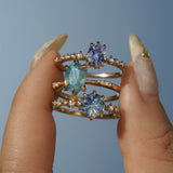 A group of three one of a kind rings by Laurie Fleming Jewellery. From top to bottom: a Nereid ring featuring an oval cut light blue purple pastel sapphire, an Ilona ring featuring an oval cut teal opalescent sapphire, and a Nereid ring with a John Dyer Starbrite cut blue sapphire centre. The rings are held between index finger and thumb and are held up against a bright blue sky.
