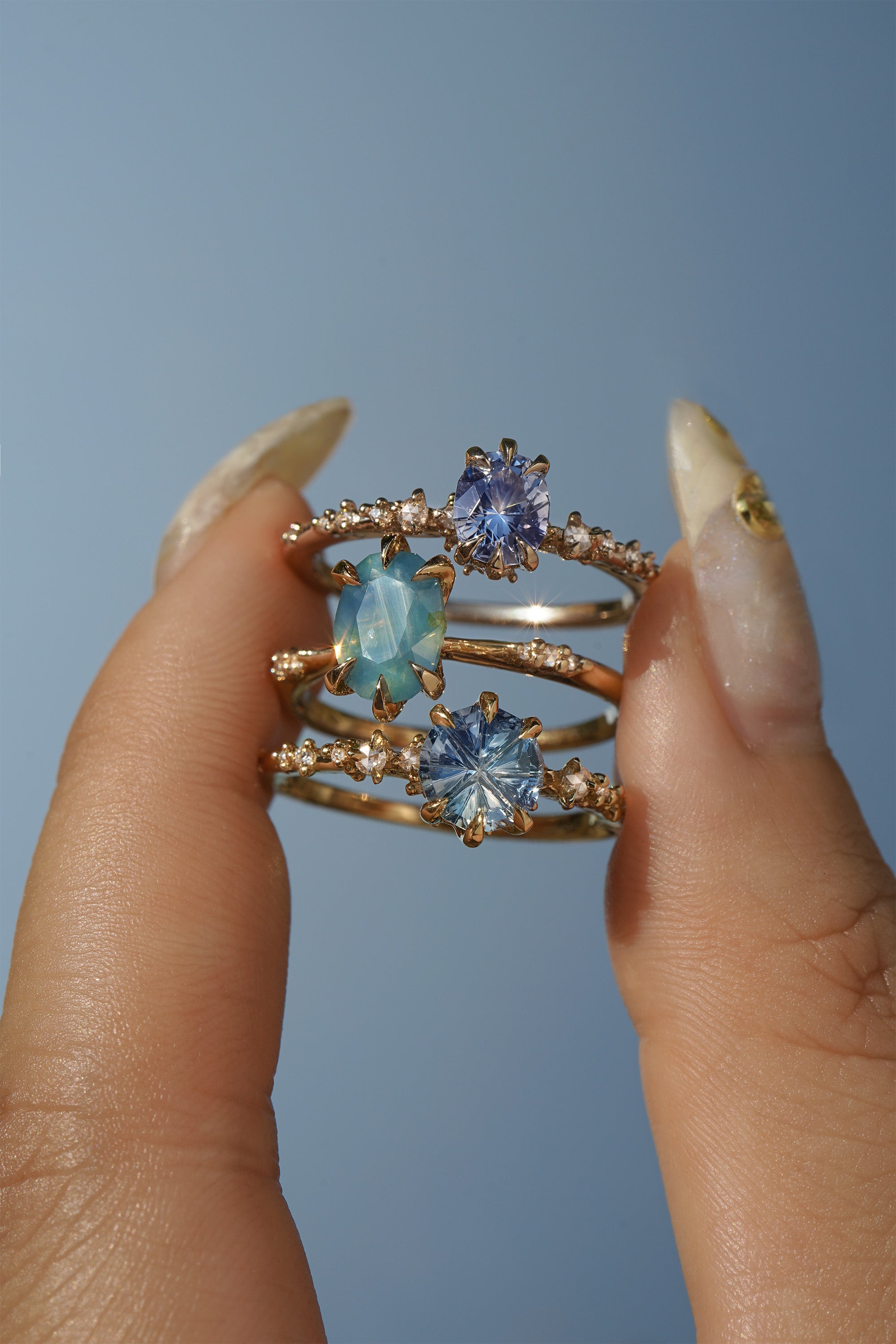 A group of three one of a kind rings by Laurie Fleming Jewellery. From top to bottom: a Nereid ring featuring an oval cut light blue purple pastel sapphire, an Ilona ring featuring an oval cut teal opalescent sapphire, and a Nereid ring with a John Dyer Starbrite cut blue sapphire centre. The rings are held between index finger and thumb and are held up against a bright blue sky.