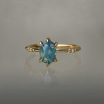 One of a kind "Ilona" ring by Laurie Fleming Jewellery featuring an oval cut opalescent turquoise teal sapphire with golden yellow sparkles. The ring has a delicate, tapered band with small diamond accents. It is in front of a medium grey background.