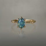 One of a kind "Ilona" ring by Laurie Fleming Jewellery featuring an oval cut opalescent turquoise teal sapphire with golden yellow sparkles. The ring has a delicate, tapered band with small diamond accents. It is in front of a medium grey background.