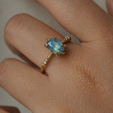 One of a kind "Ilona" ring by Laurie Fleming Jewellery featuring an oval cut opalescent turquoise teal sapphire with golden yellow sparkles. The ring has a delicate, tapered band with small diamond accents. The ring is worn on a hand.