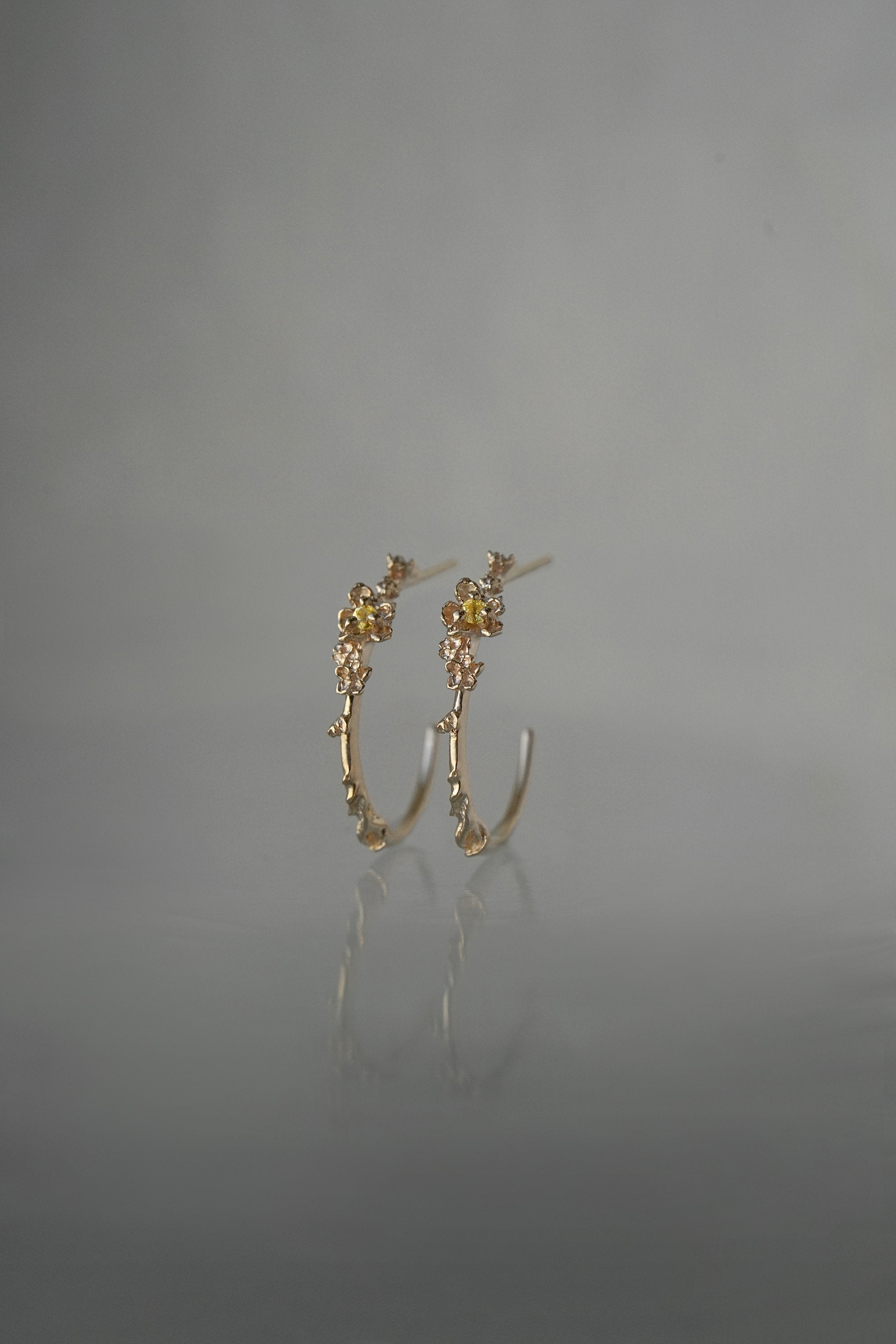 A pair of Laurie Fleming Jewellery "Large Buttercup Hoop" earrings, featuring petite hand-carved buttercup flowers with a yellow sapphire centre. The earrings are on a medium grey background.