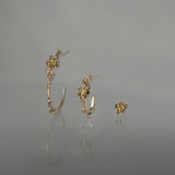 A line-up of three Laurie Fleming Jewellery earrings over a medium grey background. On the left, a large buttercup hoop. In the middle, a small buttercup hoop. On the right, a buttercup stud earring. All three earrings feature hand-carved petite buttercup flower blossoms with yellow sapphire centres.