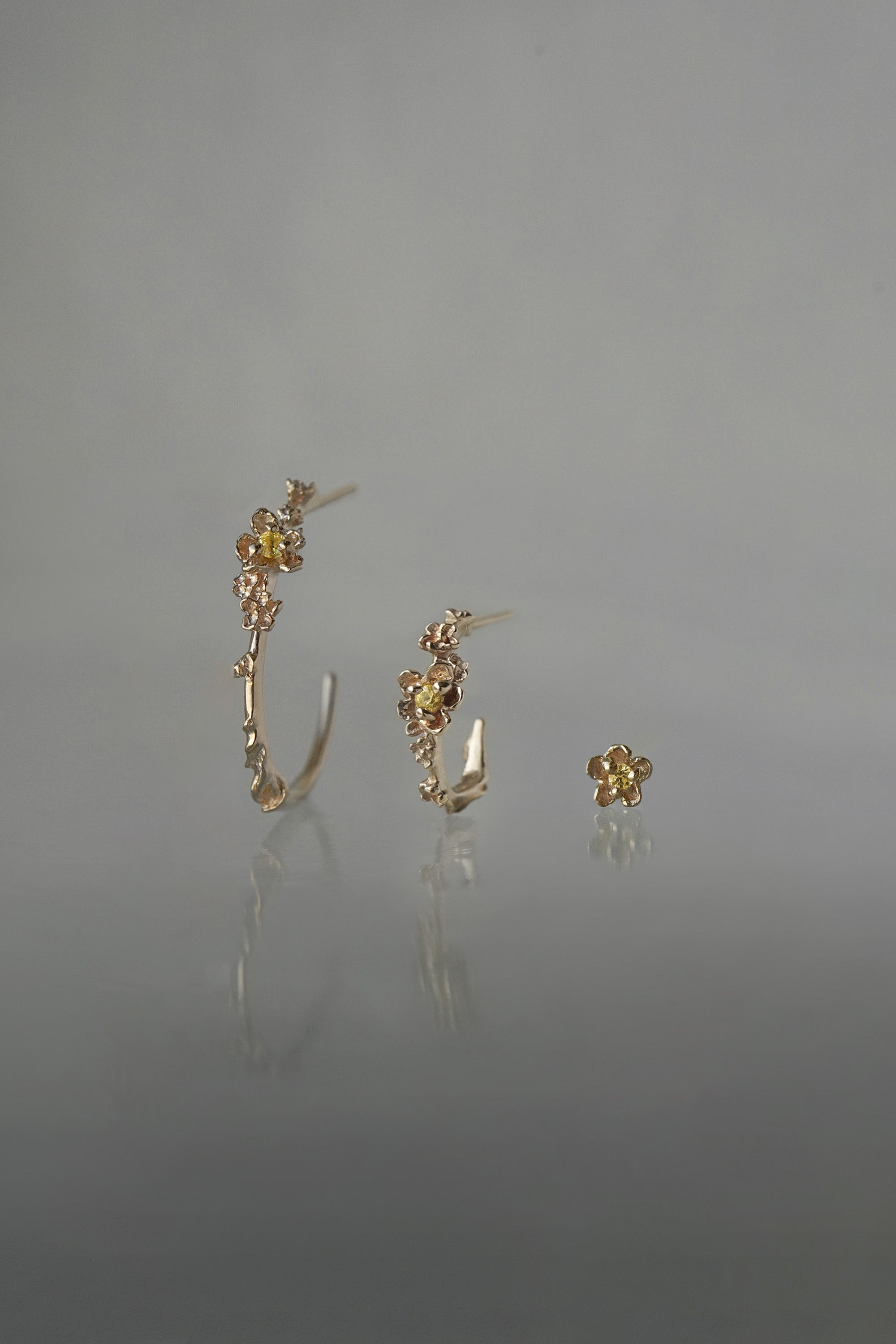 A line-up of three Laurie Fleming Jewellery earrings over a medium grey background. On the left, a large buttercup hoop. In the middle, a small buttercup hoop. On the right, a buttercup stud earring. All three earrings feature hand-carved petite buttercup flower blossoms with yellow sapphire centres.