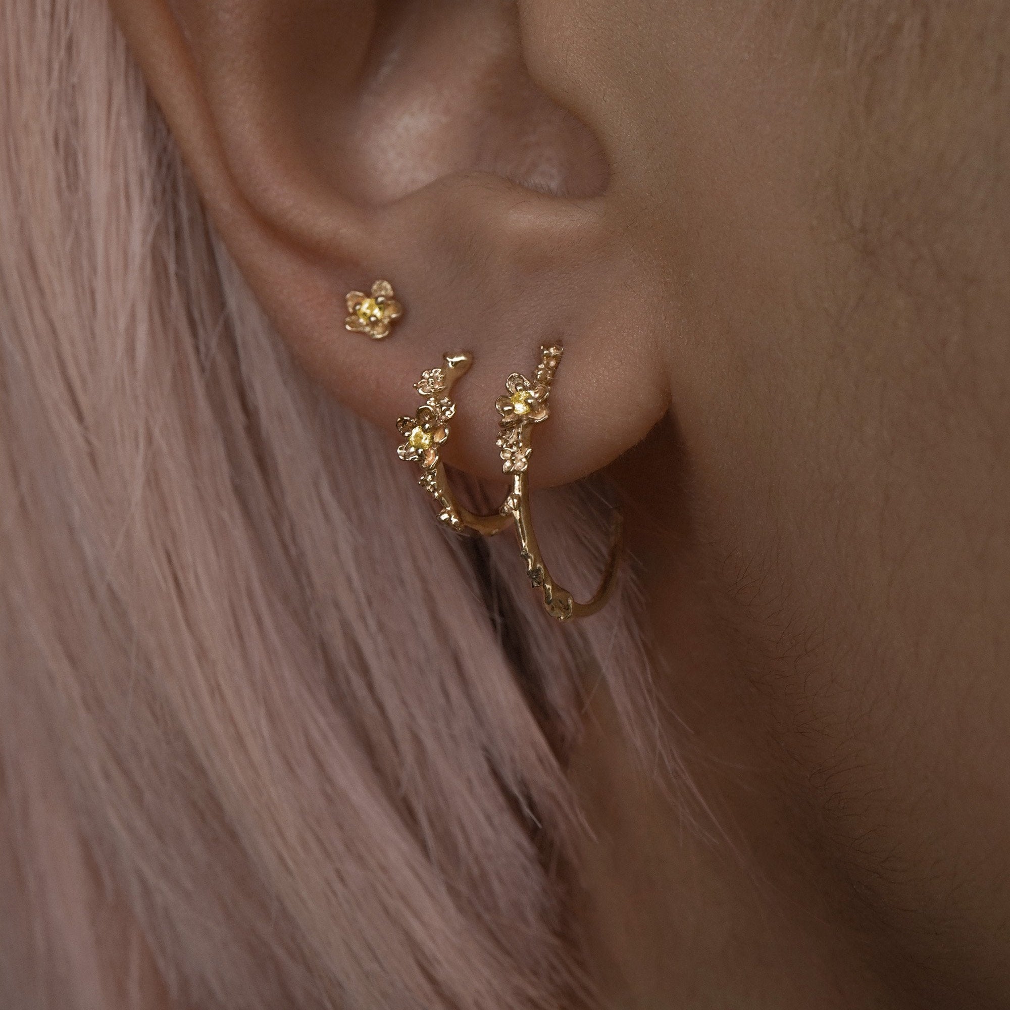 A close up of an ear wearing three Laurie Fleming Jewellery earrings. In the first hole, a large buttercup hoop. The second hole has a small buttercup hoop. The third hole has a buttercup earring. All three earrings feature hand-carved petite buttercup flower blossoms with yellow sapphire centres.