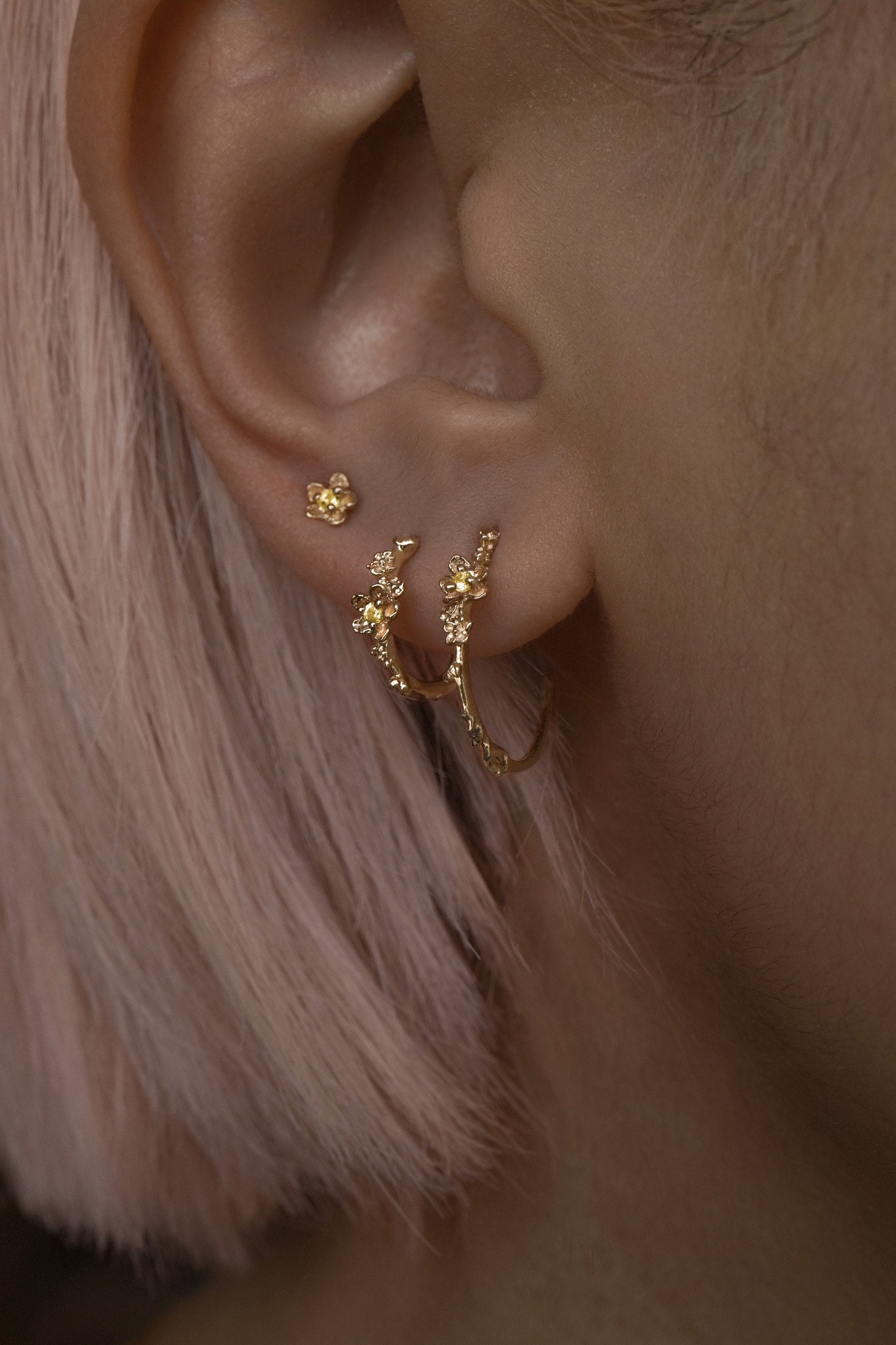 A close up of an ear wearing three Laurie Fleming Jewellery earrings. In the first hole, a large buttercup hoop. The second hole has a small buttercup hoop. The third hole has a buttercup earring. All three earrings feature hand-carved petite buttercup flower blossoms with yellow sapphire centres.