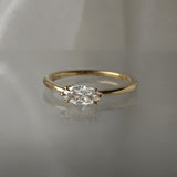 A one of a kind "Aurelia" style engagement ring by Laurie Fleming Jewellery, featuring a unique "moval" (marquise/oval hybrid) cut diamond set East-West, on a gently tapered and rounded solid gold band. The ring is situated against a medium grey background.