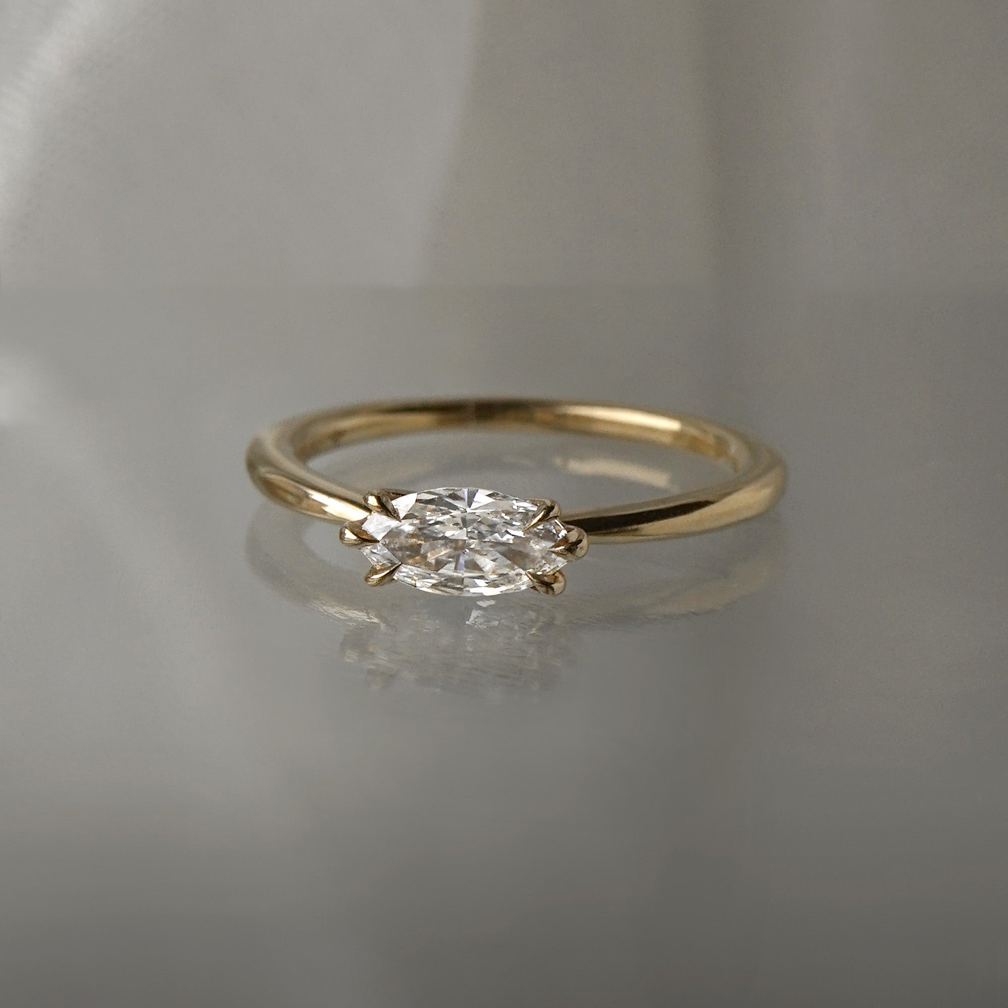 A one of a kind "Aurelia" style engagement ring by Laurie Fleming Jewellery, featuring a unique "moval" (marquise/oval hybrid) cut diamond set East-West, on a gently tapered and rounded solid gold band. The ring is situated against a medium grey background.