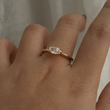 A one of a kind "Aurelia" style engagement ring by Laurie Fleming Jewellery, featuring a unique "moval" (marquise/oval hybrid) cut diamond set East-West, on a gently tapered and rounded solid gold band. The ring is worn on a hand that is posed in front of some draped gossamer silk fabric.