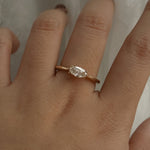 A one of a kind "Aurelia" style engagement ring by Laurie Fleming Jewellery, featuring a unique "moval" (marquise/oval hybrid) cut diamond set East-West, on a gently tapered and rounded solid gold band. The ring is worn on a hand that is posed in front of some draped gossamer silk fabric.