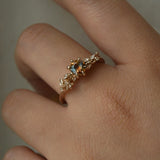 A hand wearing a one of a kind "Asai Garden" style engagement ring by Laurie Fleming Jewellery in front of a grey background. The ring features a round orange-blue "Kaleidoscope" Montana sapphire centre. The band features hand carved cherry blossom flowers with brilliant diamonds.