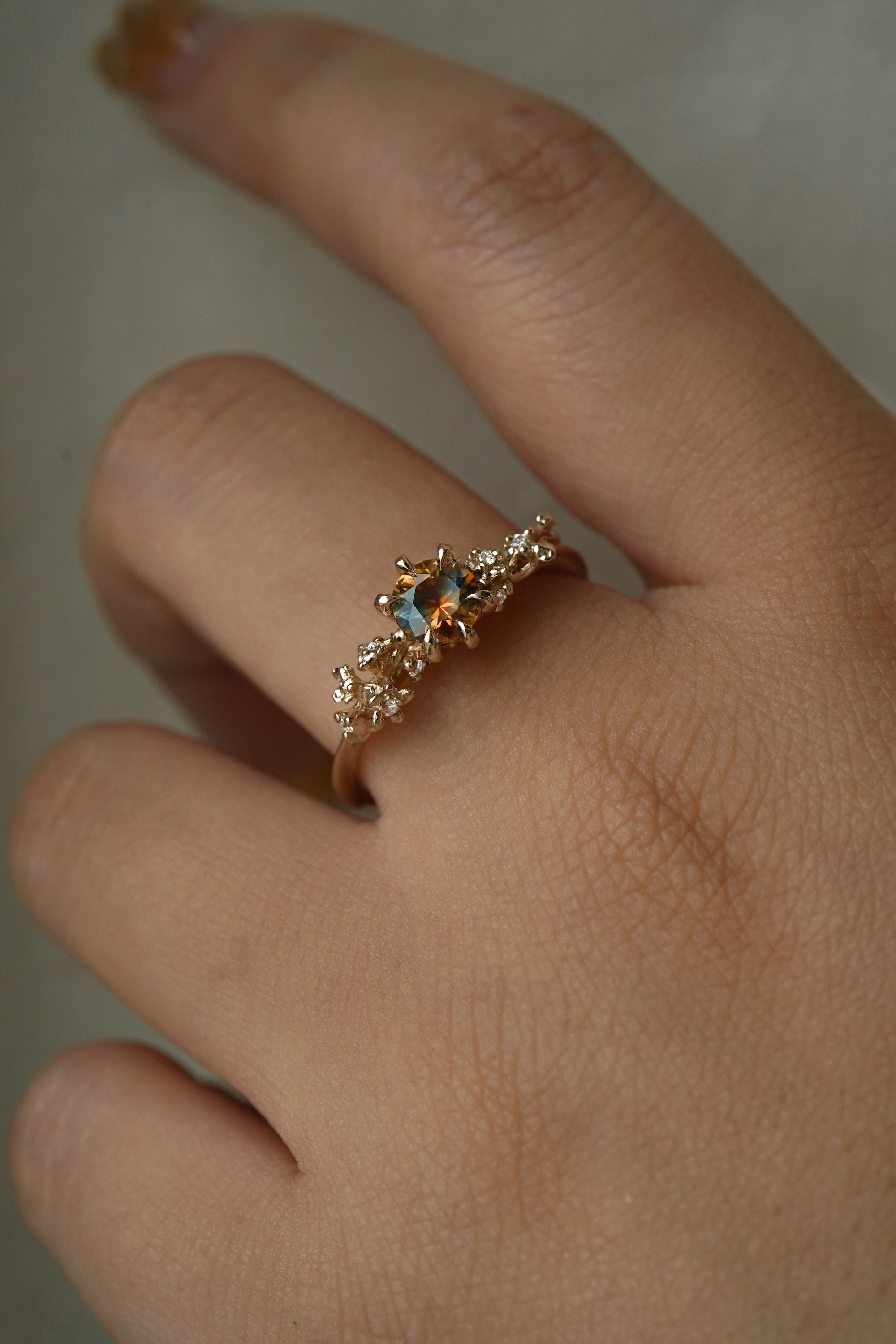 A hand wearing a one of a kind "Asai Garden" style engagement ring by Laurie Fleming Jewellery in front of a grey background. The ring features a round orange-blue "Kaleidoscope" Montana sapphire centre. The band features hand carved cherry blossom flowers with brilliant diamonds.
