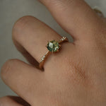 Hand wearing a dainty yellow gold Ilona style engagement ring on a grey background, with a rose cut green sapphire and diamonds on the band