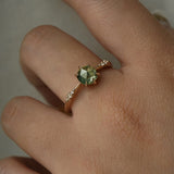 Hand wearing a dainty yellow gold Ilona style engagement ring on a grey background, with a rose cut green sapphire and diamonds on the band