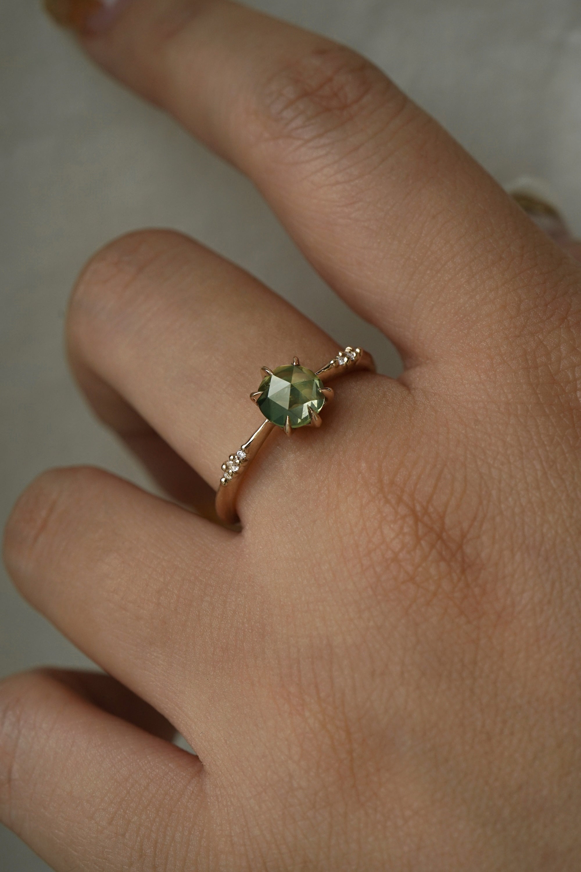 Hand wearing a dainty yellow gold Ilona style engagement ring on a grey background, with a rose cut green sapphire and diamonds on the band