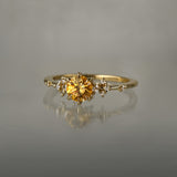 A one of a kind 18k yellow gold Water Lily engagement ring by Laurie Fleming Jewellery with a bright golden yellow round Montana sapphire centre with clusters of diamonds on the band. The ring is situated on a medium grey background.