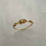 A stock photo of the solid gold "Pansy Signet" ring by Laurie Fleming Jewellery. The ring features hand-carved delicate petals encircling the band, with a tiny pansy flower at the centre, which has a customizable centre stone (pictured with a diamond, available in all birthstones). The ring is against a medium/light grey background.