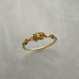 A stock photo of the solid gold "Pansy Signet" ring by Laurie Fleming Jewellery. The ring features hand-carved delicate petals encircling the band, with a tiny pansy flower at the centre, which has a customizable centre stone (pictured with a diamond, available in all birthstones). The ring is against a medium/light grey background.
