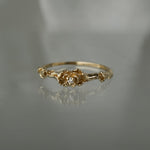 A stock photo of the solid gold "Pansy Signet" ring by Laurie Fleming Jewellery. The ring features hand-carved delicate petals encircling the band, with a tiny pansy flower at the centre, which has a customizable centre stone (pictured with a diamond, available in all birthstones). The ring is laying down on a mirror against a medium/light grey background; its reflection below it is faint.