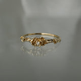A stock photo of the solid gold "Pansy Signet" ring by Laurie Fleming Jewellery. The ring features hand-carved delicate petals encircling the band, with a tiny pansy flower at the centre, which has a customizable centre stone (pictured with a diamond, available in all birthstones). The ring is laying down on a mirror against a medium/light grey background; its reflection below it is faint.