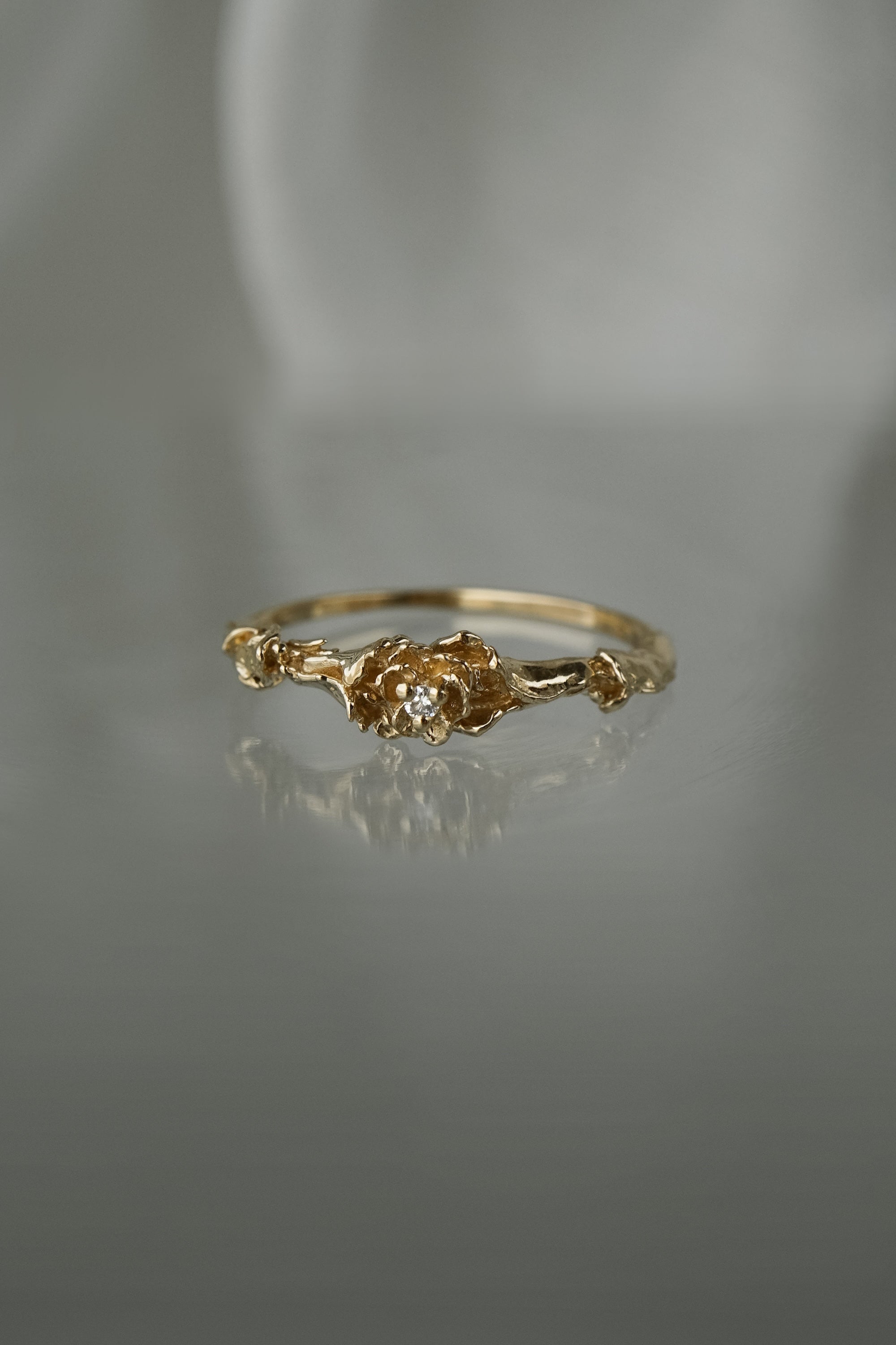 A stock photo of the solid gold "Pansy Signet" ring by Laurie Fleming Jewellery. The ring features hand-carved delicate petals encircling the band, with a tiny pansy flower at the centre, which has a customizable centre stone (pictured with a diamond, available in all birthstones). The ring is laying down on a mirror against a medium/light grey background; its reflection below it is faint.