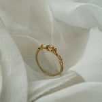 A stock photo of the solid gold "Pansy Signet" ring by Laurie Fleming Jewellery. The ring features hand-carved delicate petals encircling the band, with a tiny pansy flower at the centre, which has a customizable centre stone (pictured with a diamond, available in all birthstones). The ring is standing on its band and surrounded by ripples of raw white silk fabric.