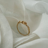 A stock photo of the solid gold "Pansy Signet" ring by Laurie Fleming Jewellery. The ring features hand-carved delicate petals encircling the band, with a tiny pansy flower at the centre, which has a customizable centre stone (pictured with a diamond, available in all birthstones). The ring is standing on its band and surrounded by ripples of raw white silk fabric.