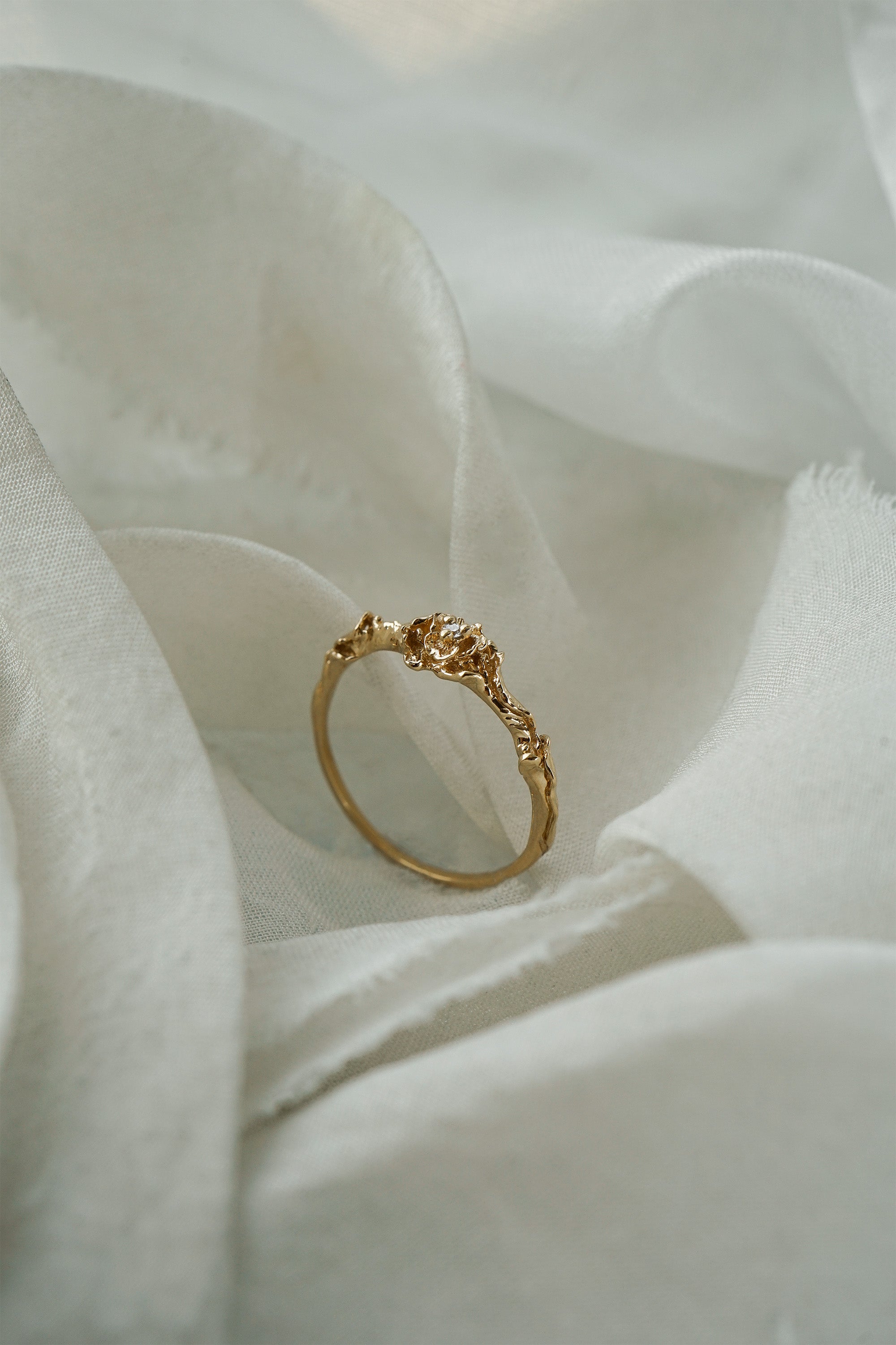 A stock photo of the solid gold "Pansy Signet" ring by Laurie Fleming Jewellery. The ring features hand-carved delicate petals encircling the band, with a tiny pansy flower at the centre, which has a customizable centre stone (pictured with a diamond, available in all birthstones). The ring is standing on its band and surrounded by ripples of raw white silk fabric.