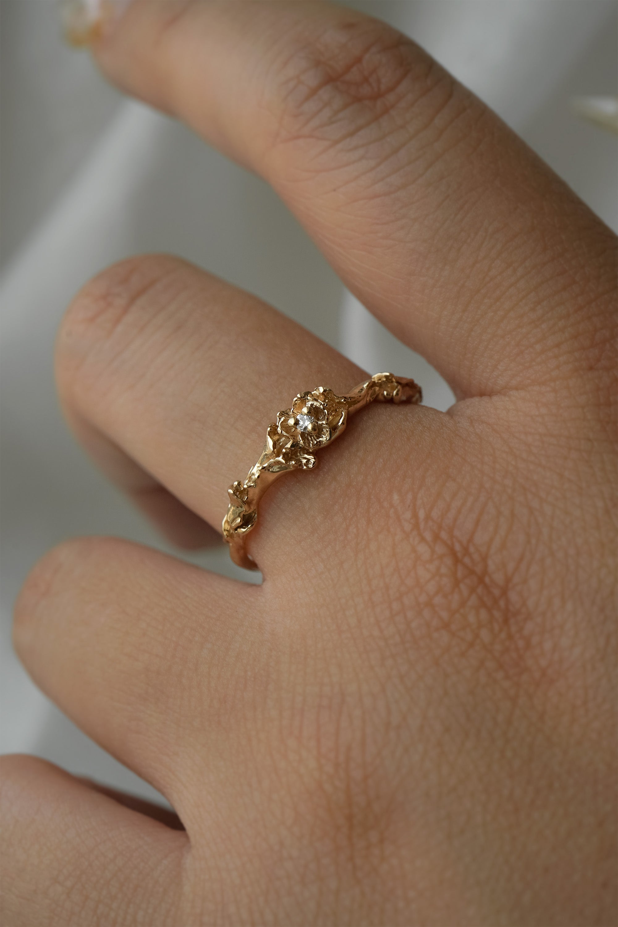 A stock photo of the solid gold "Pansy Signet" ring by Laurie Fleming Jewellery. The ring features hand-carved delicate petals encircling the band, with a tiny pansy flower at the centre, which has a customizable centre stone (pictured with a diamond, available in all birthstones). The ring is worn on a hand against a light grey background.