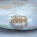 A stack of dainty Laurie Fleming Jewellery rings. The top ring is a "Pearl Water Lily Ring," a delicate solid gold stacking ring with freshwater seed pearls scattered along the band. The bottom ring is an "Asrai Garden" style ring, featuring three handcarved miniature orchid flowers with freshwater seed pearls, and lilac with pink and purple sapphires. Rings are stacked on a pearlescent oyster dish.