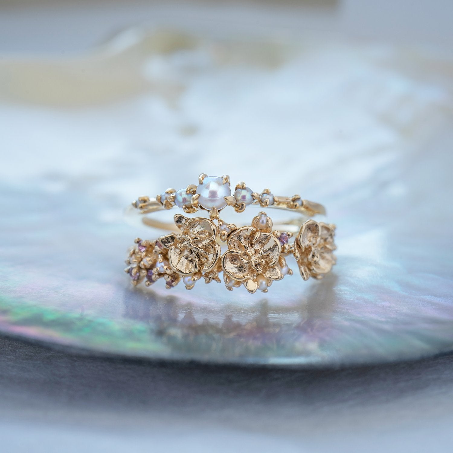 A stack of dainty Laurie Fleming Jewellery rings. The top ring is a "Pearl Water Lily Ring," a delicate solid gold stacking ring with freshwater seed pearls scattered along the band. The bottom ring is an "Asrai Garden" style ring, featuring three handcarved miniature orchid flowers with freshwater seed pearls, and lilac with pink and purple sapphires. Rings are stacked on a pearlescent oyster dish.
