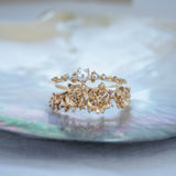 A stack of dainty Laurie Fleming Jewellery rings. The top ring is a "Pearl Water Lily Ring," a delicate solid gold stacking ring with freshwater seed pearls scattered along the band. The bottom ring is an "Asrai Garden" style ring, featuring three handcarved miniature orchid flowers with freshwater seed pearls, and lilac with pink and purple sapphires. Rings are stacked on a pearlescent oyster dish.