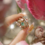 Four one of a kind engagement rings by Laurie Fleming Jewellery are held between index finger and thumb. In the foreground and background, flowers are partially visible, with the rings clearly focused in the centre. There is a Wild Iris three stone ring with a colour change sapphire centre, an Ilona ring with a seafoam green sapphire centre, a Nereid ring with a rose cut pink sapphire centre set in the middle of a blooming golden peony, and a Daphne ring with a pale rosewater pink sapphire centre.