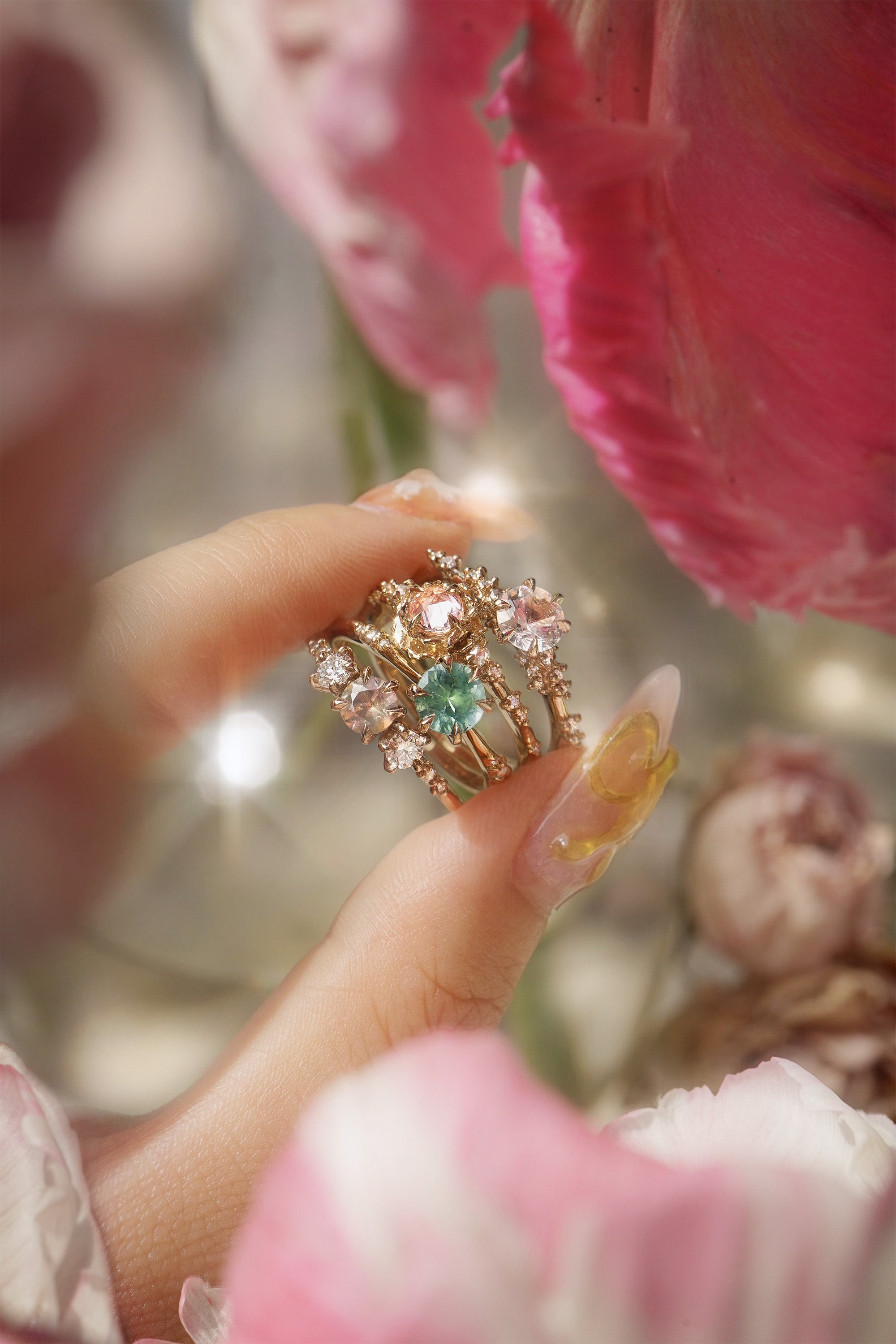 Four one of a kind engagement rings by Laurie Fleming Jewellery are held between index finger and thumb. In the foreground and background, flowers are partially visible, with the rings clearly focused in the centre. There is a Wild Iris three stone ring with a colour change sapphire centre, an Ilona ring with a seafoam green sapphire centre, a Nereid ring with a rose cut pink sapphire centre set in the middle of a blooming golden peony, and a Daphne ring with a pale rosewater pink sapphire centre.