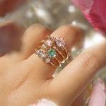 Four one of a kind engagement rings by Laurie Fleming Jewellery are worn on a hand. In the foreground and background, flowers are partially visible, with the rings clearly focused in the centre. There is a Wild Iris three stone ring with a colour change sapphire centre, an Ilona ring with a seafoam green sapphire centre, a Nereid ring with a rose cut pink sapphire centre set in the middle of a blooming golden peony, and a Daphne ring with a pale rosewater pink sapphire centre.