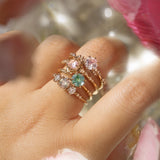 Four one of a kind engagement rings by Laurie Fleming Jewellery are worn on a hand. In the foreground and background, flowers are partially visible, with the rings clearly focused in the centre. There is a Wild Iris three stone ring with a colour change sapphire centre, an Ilona ring with a seafoam green sapphire centre, a Nereid ring with a rose cut pink sapphire centre set in the middle of a blooming golden peony, and a Daphne ring with a pale rosewater pink sapphire centre.