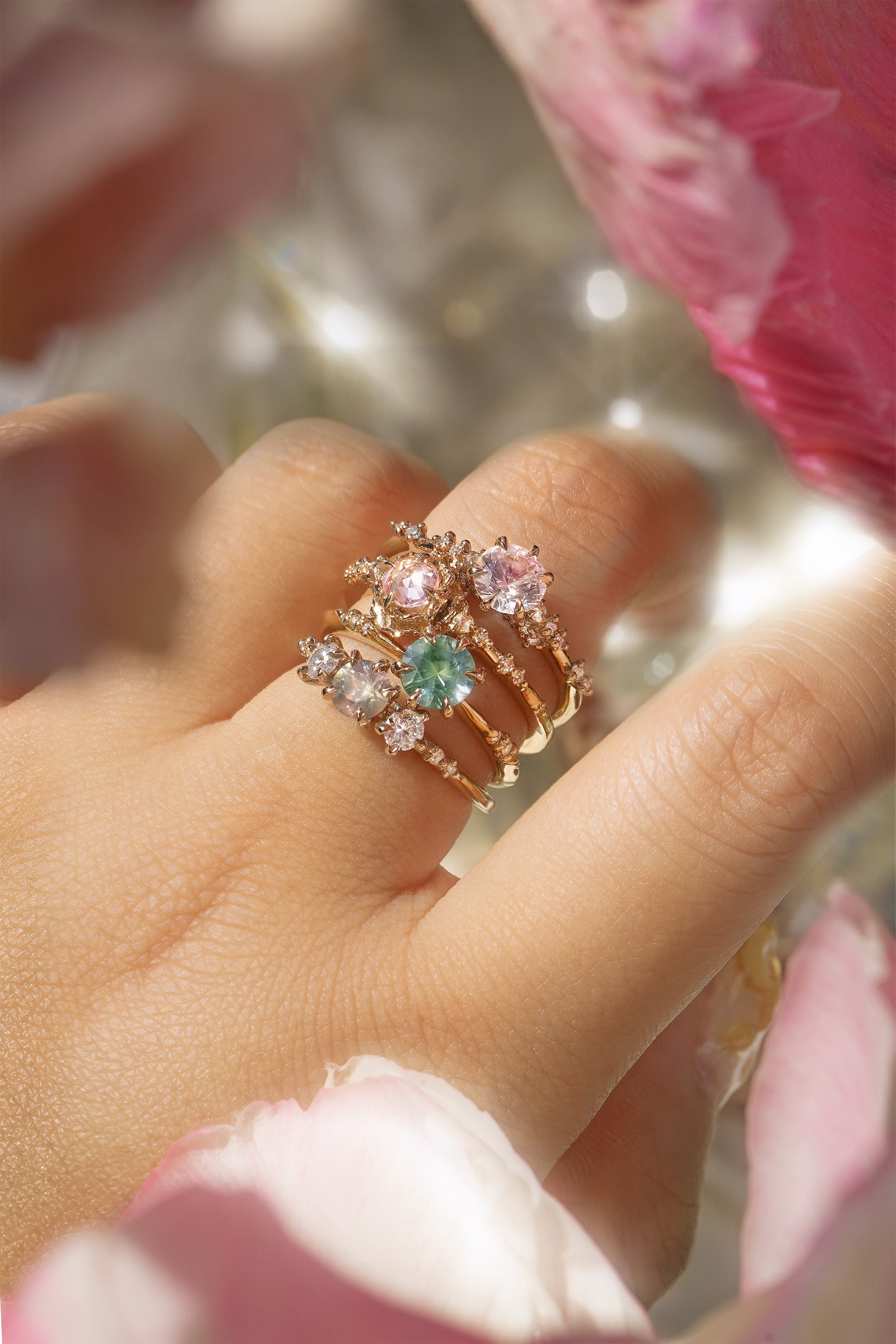 Four one of a kind engagement rings by Laurie Fleming Jewellery are worn on a hand. In the foreground and background, flowers are partially visible, with the rings clearly focused in the centre. There is a Wild Iris three stone ring with a colour change sapphire centre, an Ilona ring with a seafoam green sapphire centre, a Nereid ring with a rose cut pink sapphire centre set in the middle of a blooming golden peony, and a Daphne ring with a pale rosewater pink sapphire centre.