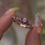 A delicate and ethereal hand-carved one of a kind "Daphne" style engagement ring by Laurie Fleming Jewellery. The ring features a round brilliant cut red peach padparascha sapphire centre, with clusters of rose and brilliant cut diamond accents on the band in 14k rose gold. The ring is held between an index finger and thumb and rotated gently from side to side. The background, which is blurred and out of focus, is of plants.