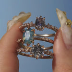 A group of three one of a kind rings by Laurie Fleming Jewellery. From top to bottom: a Nereid ring featuring an oval cut light blue purple pastel sapphire, an Ilona ring featuring an oval cut teal opalescent sapphire, and a Nereid ring with a John Dyer Starbrite cut blue sapphire centre. The rings are held between index finger and thumb and are held up and turned side to side against a bright blue sky.