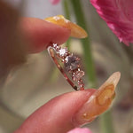 A one of a kind "Wild Iris" style engagement ring by Laurie Fleming Jewellery featuring a colour-change sapphire centre with shifting hues of blue, grey, blush, and green, flanked by a diamond on either side. Additional diamonds drip down the band. The ring is handcarved and made in solid champagne yellow gold. The ring is held between index finger and thumb and rotated from side to side, with flowers partially visible in the foreground and background.
