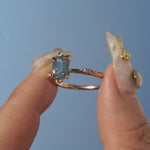 One of a kind "Ilona" ring by Laurie Fleming Jewellery featuring an oval cut opalescent turquoise teal sapphire with golden yellow sparkles. The ring has a delicate, tapered band with small diamond accents. The ring is held up between index finger and thumb and rotated from side to side, in front of a bright blue sky.