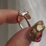 A hand holding a one of a kind "Ilona" style engagement ring between index finger and thumb, rotating it side to side. The ring features a 0.92ct natural Emerald cut diamond held by four delicately hand-carved claw shaped prongs. The band tapers towards the centre and features diamonds on the shoulders. The background is a light grey with some draped silk fabric.