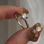 A hand holding a delicate hand-carved one of a kind "Ilona" style engagement ring by Laurie Fleming Jewellery between index finger and thumb, rotating it side to side. The ring features a 0.8ct round brilliant cut diamond, a tapered knife-edge band, and small diamond accents on the band shoulders.  The background is a light grey with some draped silk fabric.