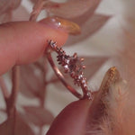 A delicate and ethereal hand-carved one of a kind "Daphne" style engagement ring by Laurie Fleming Jewellery. The ring features a rose cut pale pink sapphire centre, with clusters of rose and brilliant cut diamond accents on the band in solid 14k rose gold. The ring is held between index finger and thumb of a hand and is rotated from side to side. The background features dried light red leaves and the foreground has a fluffy plant partially visible.