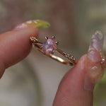 A delicate hand-carved one of a kind "Ilona" style engagement ring by Laurie Fleming Jewellery. The ring features a silky and opalescent rose cut oval pink sapphire centre, a tapered knife-edge band, and small diamond accents on the band shoulders. The ring is held between an index finger and thumb and rotated gently from side to side. The background, which is blurred and out of focus, is of plants.