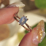 A delicate hand-carved one of a kind "Ilona" style engagement ring by Laurie Fleming Jewellery. The ring features a 0.93ct geometric oval cut silky periwinkle blue sapphire centre, a tapered knife-edge band, and small diamond accents on the band shoulders.  The ring is held between index finger and thumb and rotated slowly from side to side. In the background, there are out-of-focus plants rustling in the breeze.