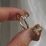 A hand holding a delicate hand-carved one of a kind "Ilona" style engagement ring by Laurie Fleming Jewellery between index finger and thumb, rotating it side to side. The ring features a 0.5ct marquise cut diamond, a tapered knife-edge band, and small diamond accents on the band shoulders.  The background is a light grey with some draped silk fabric.
