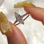 A one of a kind handcarved "Moondew" style engagement ring by Laurie Fleming Jewellery in solid 14k champagne gold. The ring features a unique moval shaped diamond (a long, marquise-oval mix) on a thoughtfully tapered plain gold band, held securely by four prongs at North, South, East, and West. The ring is held between an index finger and thumb, and rotated gently from side to side. The background is a pale gray with flecks of rainbow light.